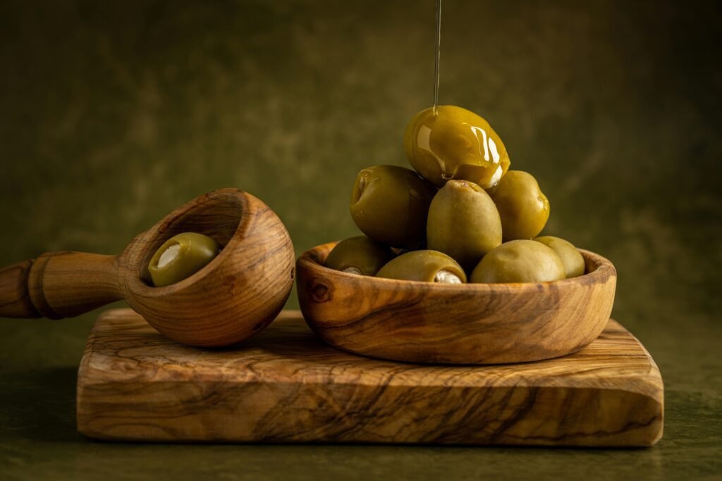 An artistic composition of green olives drizzled with oil in a wooden bowl on a rustic cutting board.